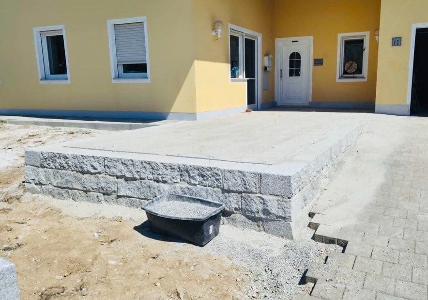 Neu gebaute Terrasse vor einem Wohnhaus, eingefasst von einer Mauer aus Granitsteinen, daneben frisch verlegtes Pflaster